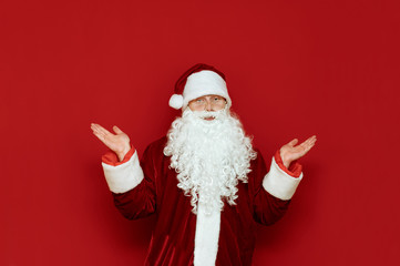 Fototapeta na wymiar Studio portrait of a funny Santa Claus on a red background, shaking his arms in confusion and looking into the camera with a bewildered face. Confused Santa isolated on red background. X-mas