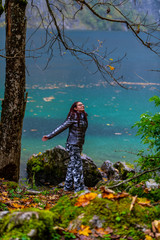 a young, red-haired girl with glasses, fashionable, sportily dressed stands in the rain, in the background an alpine lake