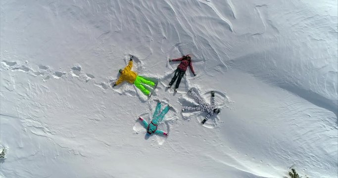 AERIAL CLOSE UP rotation: four people lie in the fresh snow and move their arms and legs, depicting a snow angel. happy two girls and two guys in bright ski suits on white snow like angels of winter.