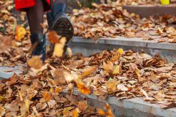 A girl in black boots and a red coat kicks yellow and red foliage strolling in the park alone on a clear autumn day during a fall. Freshness, nature and fun.