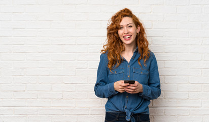 Redhead woman over white brick wall sending a message with the mobile