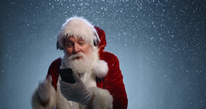 Funny Senior man in santa claus clothes and headphones is dancing to music played on his phone, isolated on blue snowy background - christmas spirit, christmas party close up 4k footage