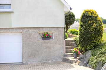 Fototapeta na wymiar Village house with garden. Entrance to the garage in a country house. Geranium flowers on the window. Gray private house with a garden and flowers