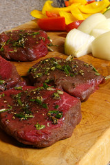 Venison steaks on wooden cutting board, spiced with garlic, rosemary and thyme. Chopped vegetables on background. Vertical image.
