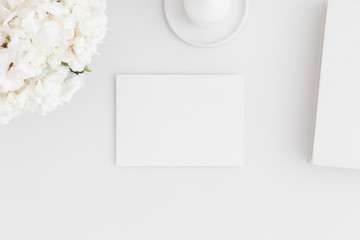 Top view of a white card mockup with workspace accessories and flowers on a white table.