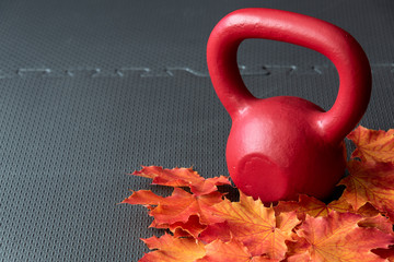 Red iron kettlebell on a black rubber gym floor, with orange and yellow maple leaves, fall fitness