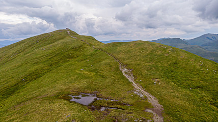 An aerial view of a grassy mountain rocky summit and mountain range in the background under a cloudy sky