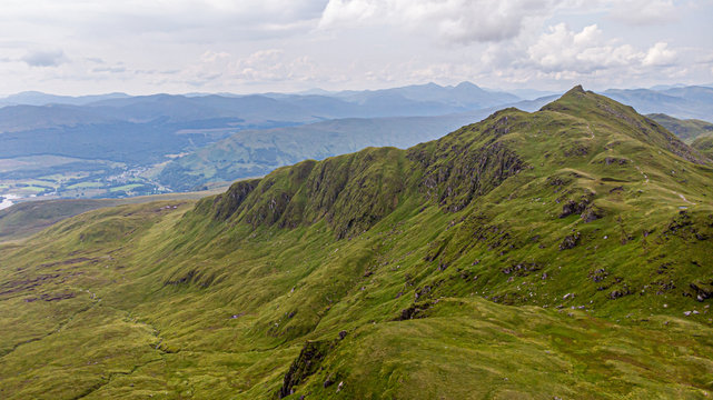 An aerial view of a grassy mountain rocky summit and mountain range in the background under a cloudy sky