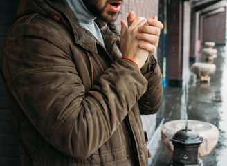 handsome man standing on the street in winter time and warming his hands. cold day. it's raining. man in jacket. Town architecture on the background. Outdoors