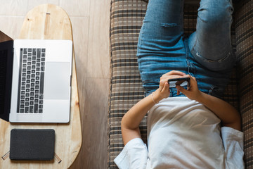 Woman use a smartphone while lying on a sofa with a laptop and ereader on a table. Studying online, freelance. Self employed or freelance girl use a phone and resting from work whith a notebook.