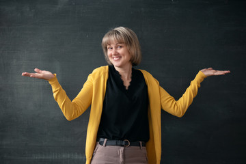 Young teacher standing near blackboard in a classroom and is shrugging her shoulders.