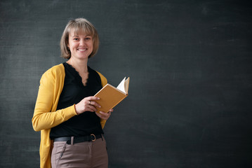 Teacher with textbook is standing near blackboard with copy space in a classroom.