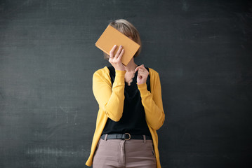 Student hiding her face behind a textbook on blackboard background.