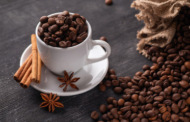 White cup full of coffee beans cinnamon bag spills out coffee stars anise star anise on a dark background