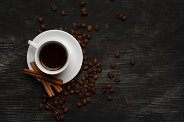 White cup with coffee cinnamon sticks on a black background with coffee beans and place for text top view