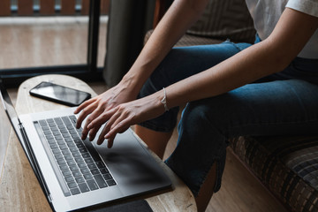 Naklejka premium Woman is sitting on a sofa and using a laptop at wooden table. Study and work online, freelance. Self employed girl is working with her notebook sitting on a couch with a phone and ereader on table.
