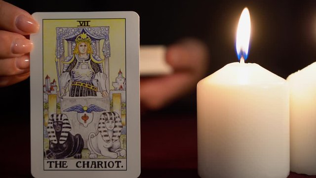 A witch is fortune teller in black mantle with manicure shows tarot card of 7 Arcana: The Chariot. White candles burns on table. Occult, esoteric, divination and wicca concept. Magician guesses.