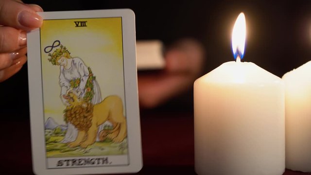 A witch is fortune teller in black mantle with manicure shows tarot card of 8 Arcana: Strength. White candles burns on table. Occult, esoteric, divination and wicca concept. Magician guesses.