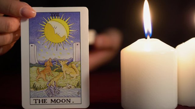 A witch is fortune teller in black mantle with manicure shows tarot card of 18 Arcana: The Moon. White candles burns on table. Occult, esoteric, divination and wicca concept. Magician guesses.
