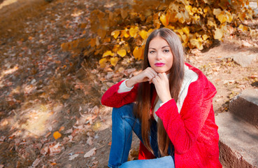 Young beautiful brunette woman with red lips and long straight hair wearing red jacket sitting outdoor in autumn park.