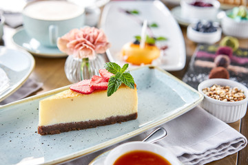 Piece of delicious cheesecake with strawberry and mint leaves on white plate. Breakfast in the cafe, morning coffee. Cappuccino and lots of desserts on the table.