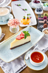 Piece of delicious cheesecake with strawberry and mint leaves on white plate. Breakfast in the cafe, morning coffee. Cappuccino and lots of desserts on the table.