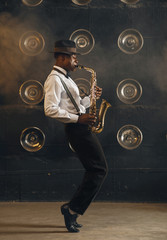 Black jazzman in hat plays the saxophone on stage - obrazy, fototapety, plakaty