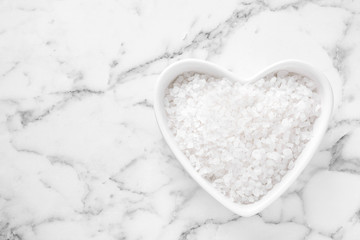 Heart shaped bowl of white sea salt on marble table, top view with space for text. Spa treatment
