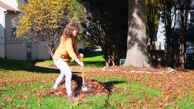 Young woman homeowner in garden yard backyard raking collecting of dry autumn foliage oak leaves doing chore with rake in sunny fall
