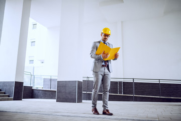 Hardworking architect with a yellow protective helmet on his head and in his grey suit standing outdoor and looking at his folder