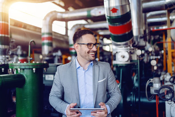 Smiling caucasian supervisor in suit and with eyeglasses holding tablet and looking away while standing in energy plant. © Dusan Petkovic