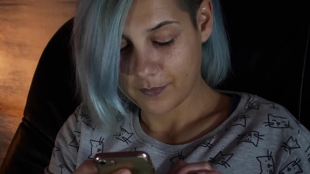 Young woman sits in chair and surfs the internet on her smartphone; with partially shaved head.