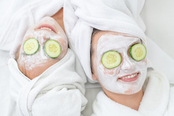 Mother and daughter in white bathrobes and with towels on their heads applying pieces of cucumber to their eyes