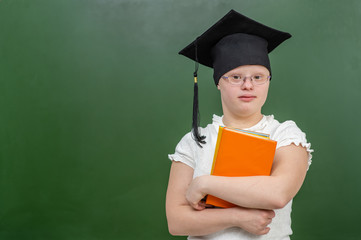 Smiling girl with Down Syndrome wearing a graduation cap near empty a chalkboard and holds books in her hands. Empty space for text