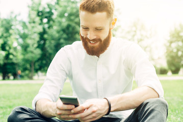 Young red-haired guy with a beard in a white shirt uses a smartphone while sitting outdoors on a sunny day