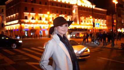 Stylish young woman in an elegant beige coat uses a smartphone standing by the road, on the street of the city at night.