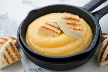 Close-up of a frying pan with polenta and slices of grilled cheese, studio shot over white concrete background