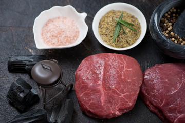 Raw marbled beefsteaks over dark brown stone background with condiments, charcoal and decorative grill, studio shot