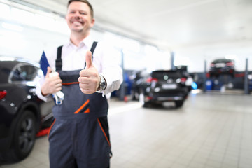 Focus on hand of happy engineer man showing thumb up and standing in modern car maintenance garage with automobiles. Machinery repairman concept. Blurred background