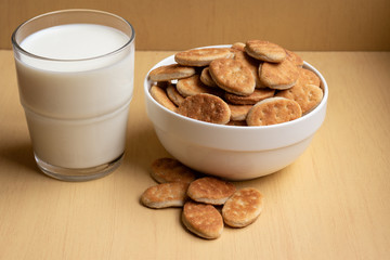 Fat cookies in a bowl, glass cup with milk, light brown wooden background.