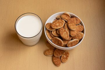 Fat cookies in a bowl, glass cup with milk, light brown wooden background.