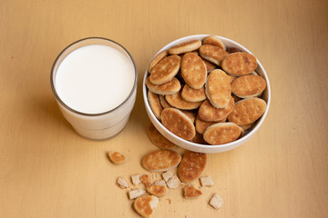 Fat cookies in a bowl, glass cup with milk, light brown wooden background.
