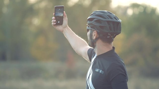 Cyclist Taking Selfie On Mobile Phone.Man Takes Picture On Phone After Cycling.Selfie Cyclist On Helmet Taking Photo With Smartphone.Athlete Answer To Video Call.Triathlete Rest On Nature With Bicycle