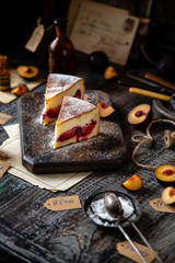 two slices of baked cheesecake or cottage cheese casserole on shortcrust with plums and powdered sugar stands on wooden board on rustic table with plums, old bottles opposite concrete wall