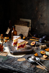 two slices of baked cheesecake or cottage cheese casserole on shortcrust with plums and powdered sugar stands on wooden board on rustic table with plums, old bottles opposite concrete wall