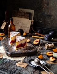 Fototapeta na wymiar two slices of baked cheesecake or cottage cheese casserole on shortcrust with plums and powdered sugar stands on wooden board on rustic table with plums, old bottles opposite concrete wall