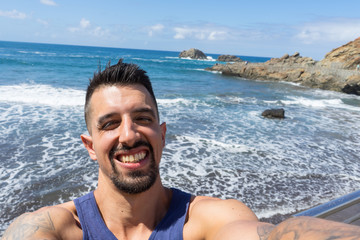 Charming bearded latino sharing selfie in social media in the beach with close eyes because of the sun. Tan Fit muscle surfer with tattoo in the arm showing teeth, charming and smiling in the ocean
