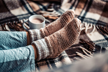 Cozy woman in knitted winter warm socks and in pajamas resting on checkered plaid blanket with a cup of hot cocoa at home in winter time. Cozy time and winter drinks. Top view