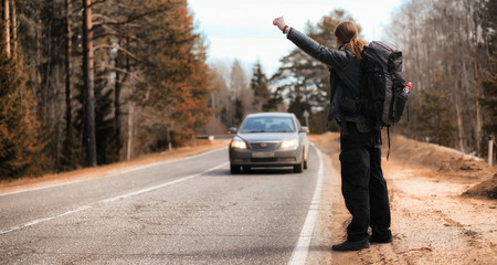 A young man is hitchhiking around the country. The man is trying to catch a passing car for traveling. The man with the backpack went hitchhiking to the south.