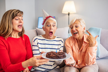 Senior woman blow on birthday cake during a birthday party. Group of seniors celebrating a birthday at home. Senior Woman Blows Out Birthday Cake Candles with females friends.